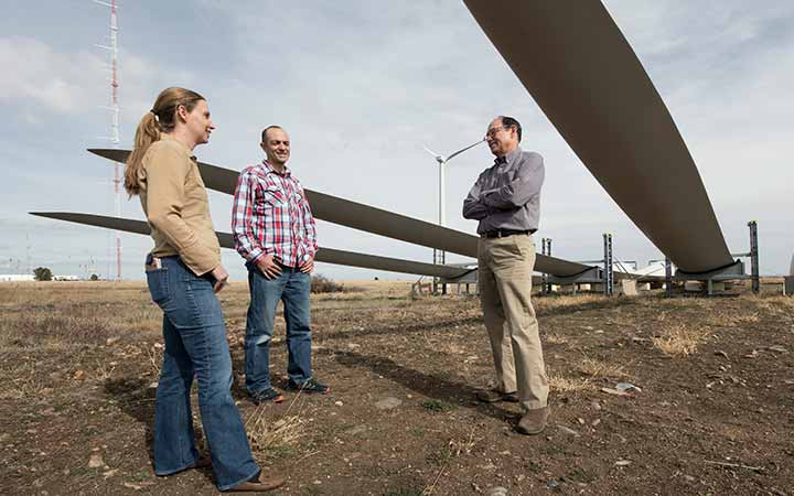 research-areas-54078 Three software co-authors stand underneath large wind turbine blades in a field outside.