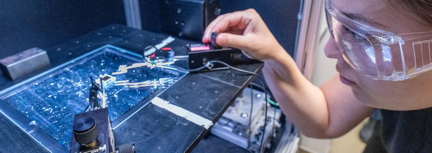81474-hero Researcher in safety goggles operating a probe station over an illuminated microfluidic chip in a laboratory.
